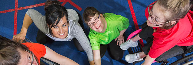 Overhead view of a group of special needs teenagers and young women with instructor, showing team spirit  They are sitting in a circle on a gym floor, hands in the center  Several of the girls have downs syndrome and two are in wheelchairs 