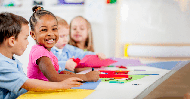 A multi-ethnic group of preschool age students drawing with crayons in their classroom at school - little girl is smiling and looking at the camera 